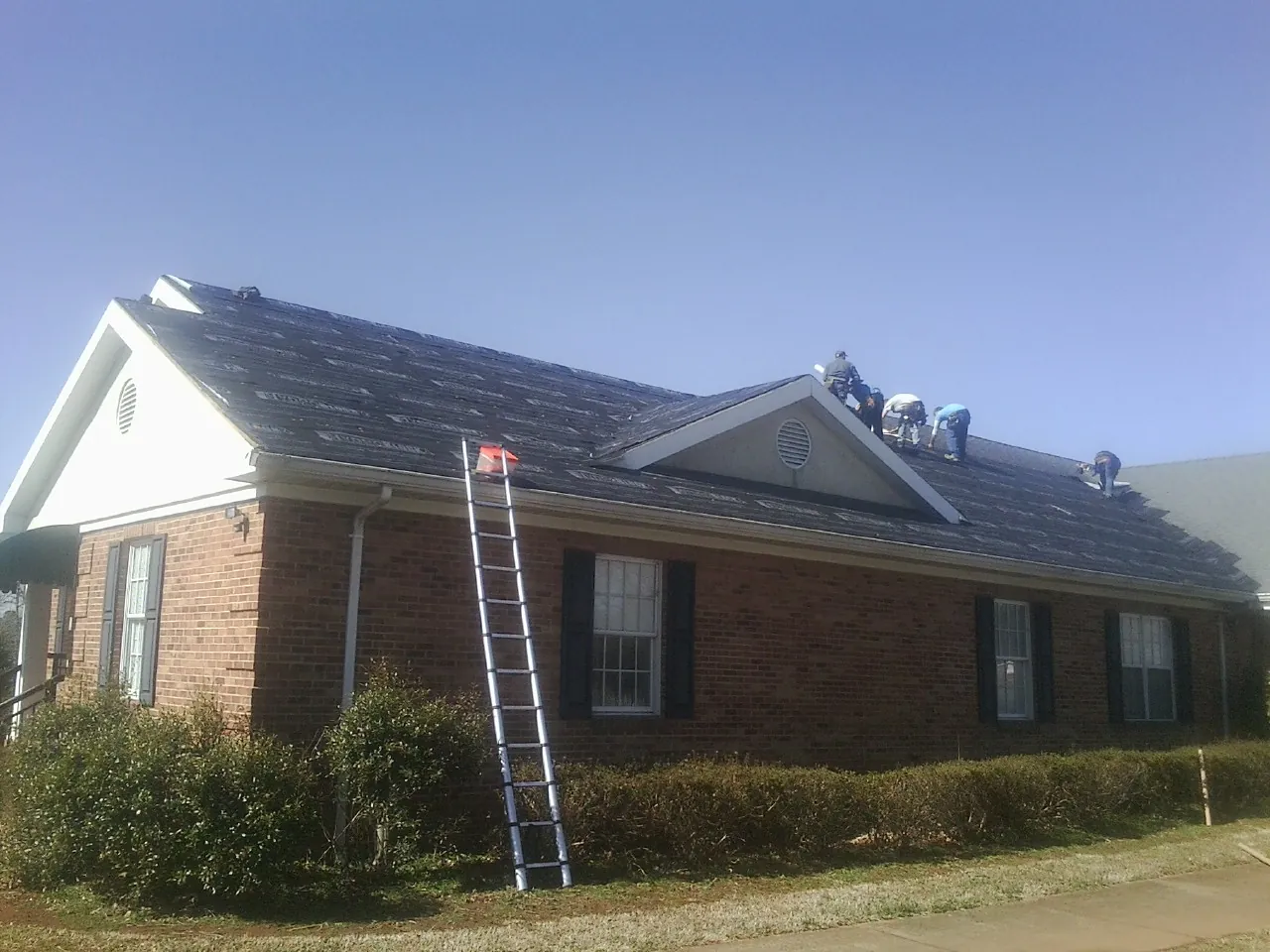 Crew tearing shingles off a brick ranch roof with a ladder set — Metal Roof Repair work by our crew in Saint Edward
