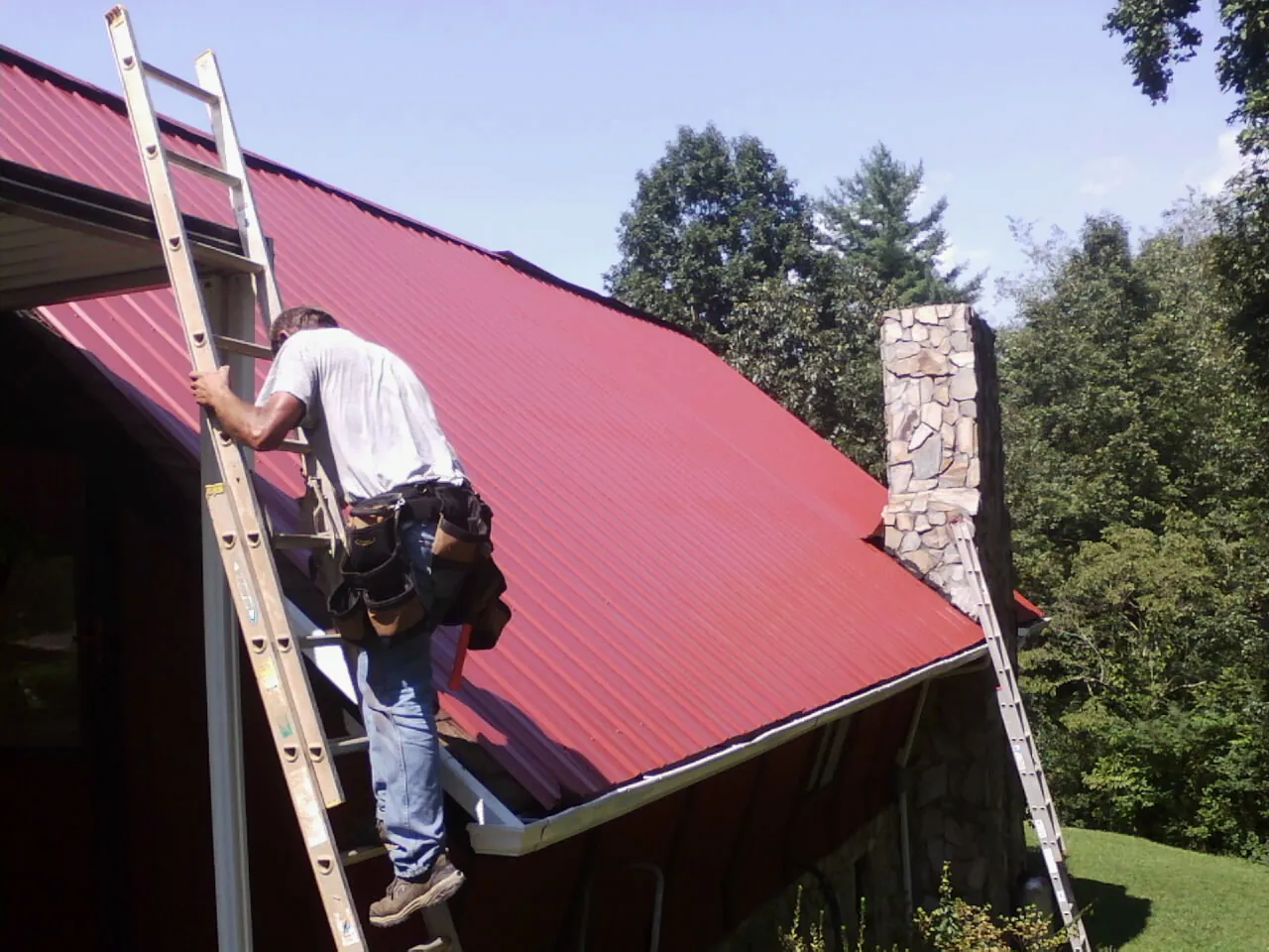 Roofer on a ladder beside a red metal roof and stone chimney — Soffit & Fascia Repair by Roofing Brothers in Saint Edward